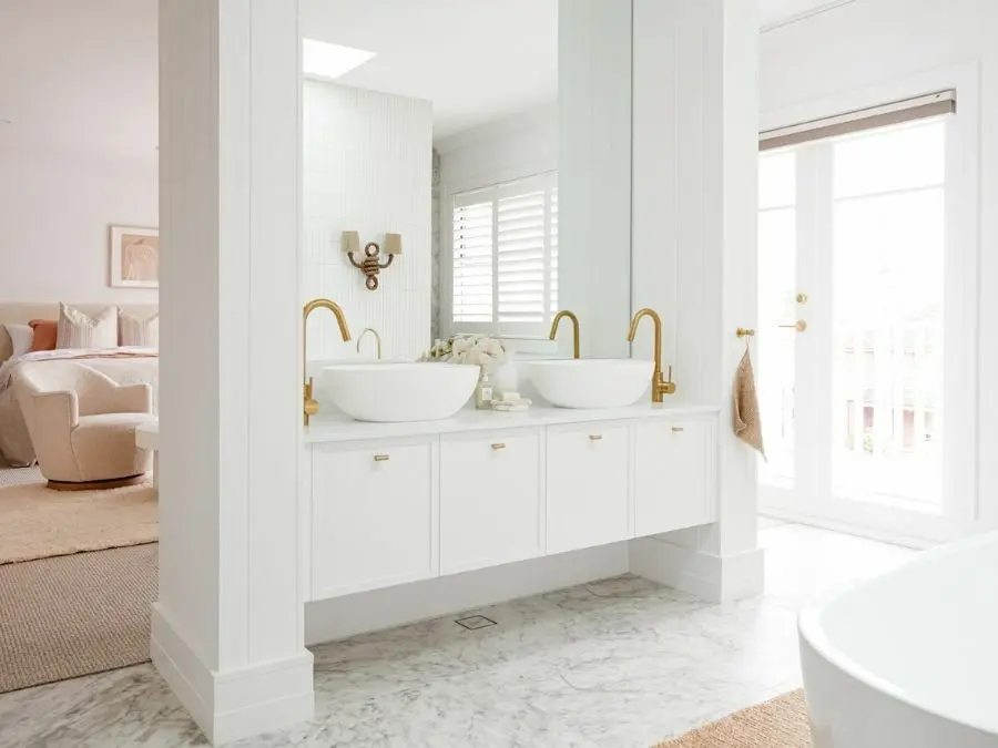 Bright white bathroom with dual vessel sinks, gold fixtures, marble floors, and a view into an adjacent bedroom.