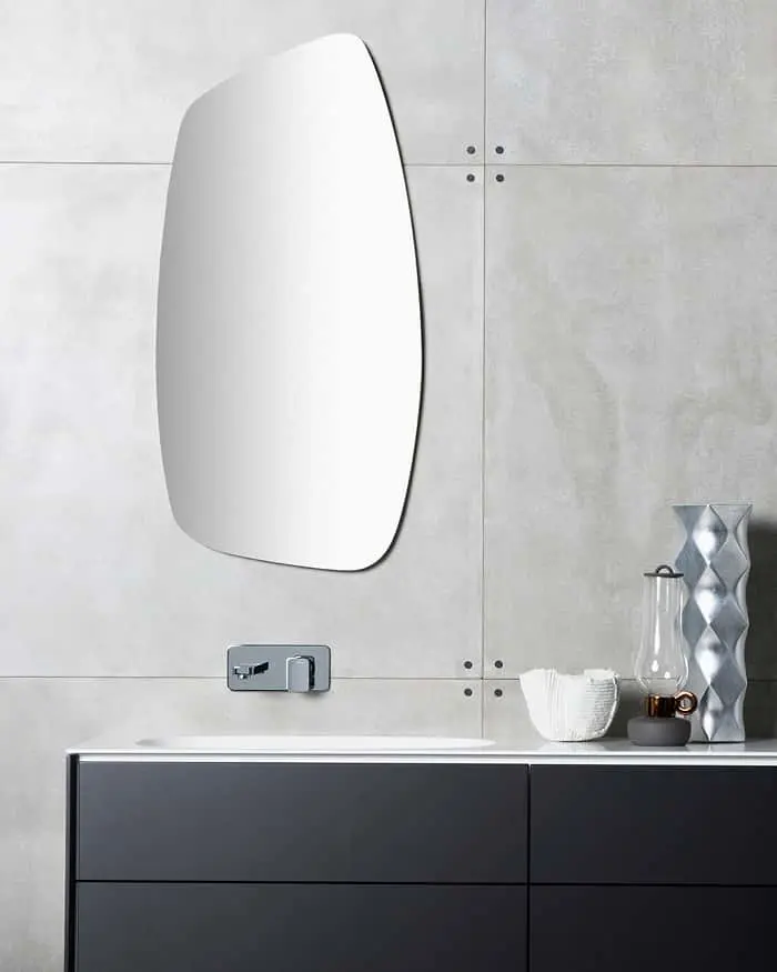 Modern bathroom with frameless oval mirror above dark vanity cabinet, metallic faucet, and decorative vases on light gray walls.
