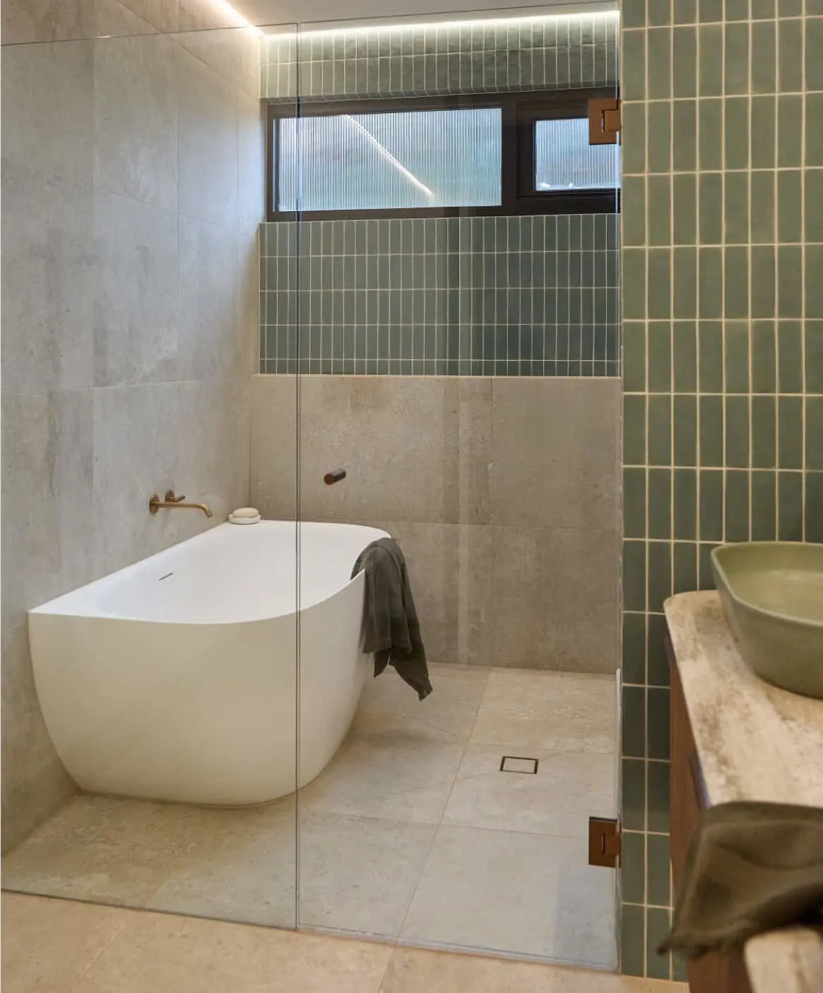 Modern bathroom with white freestanding tub, green vertical tiles, glass shower partition, and window above the bath area.