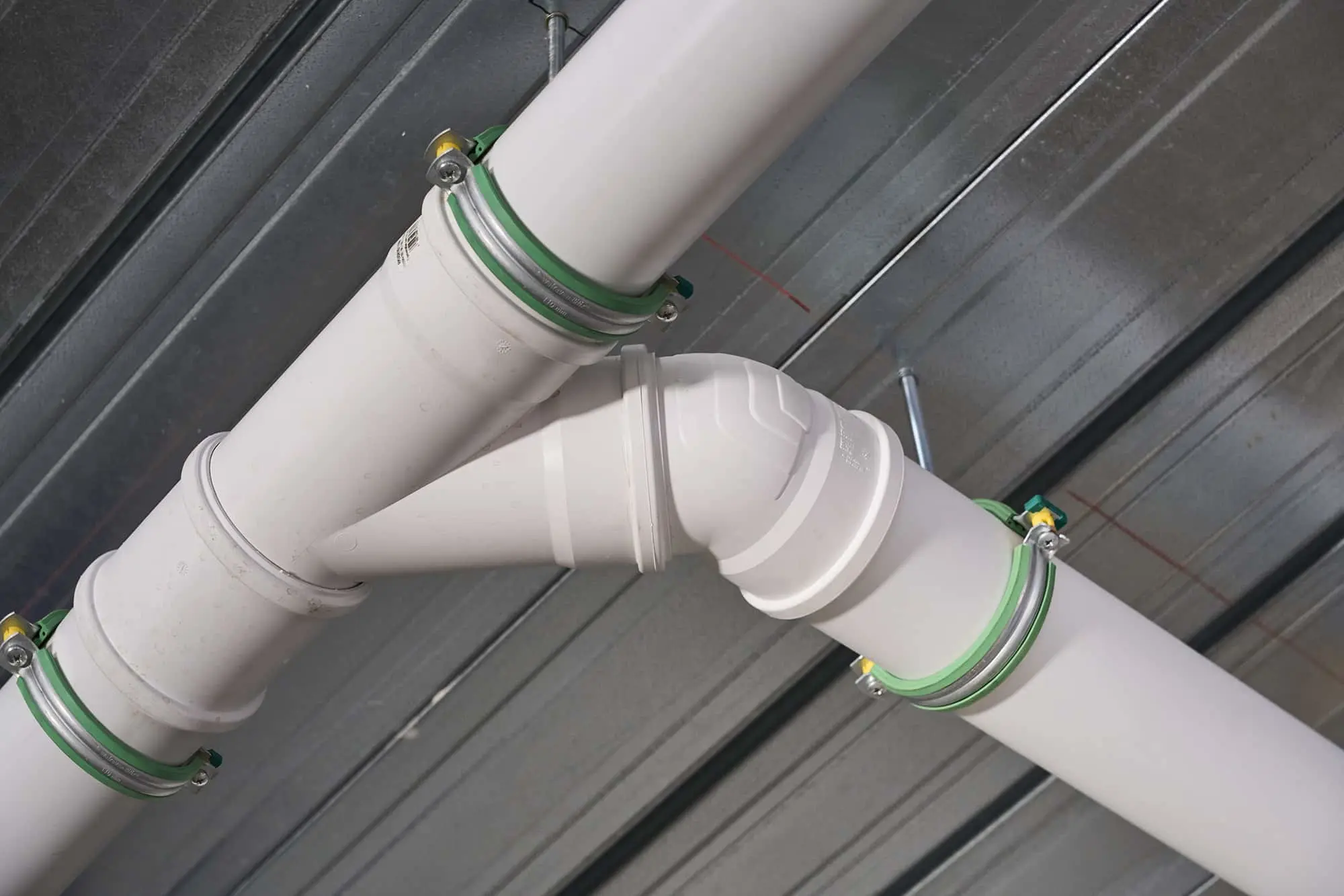 White PVC plumbing pipes with an elbow joint mounted to a metal ceiling with green and yellow clamps.