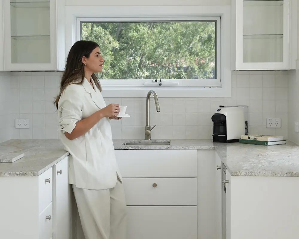 Person in white outfit holding coffee cup in modern white kitchen with coffee machine and window view.