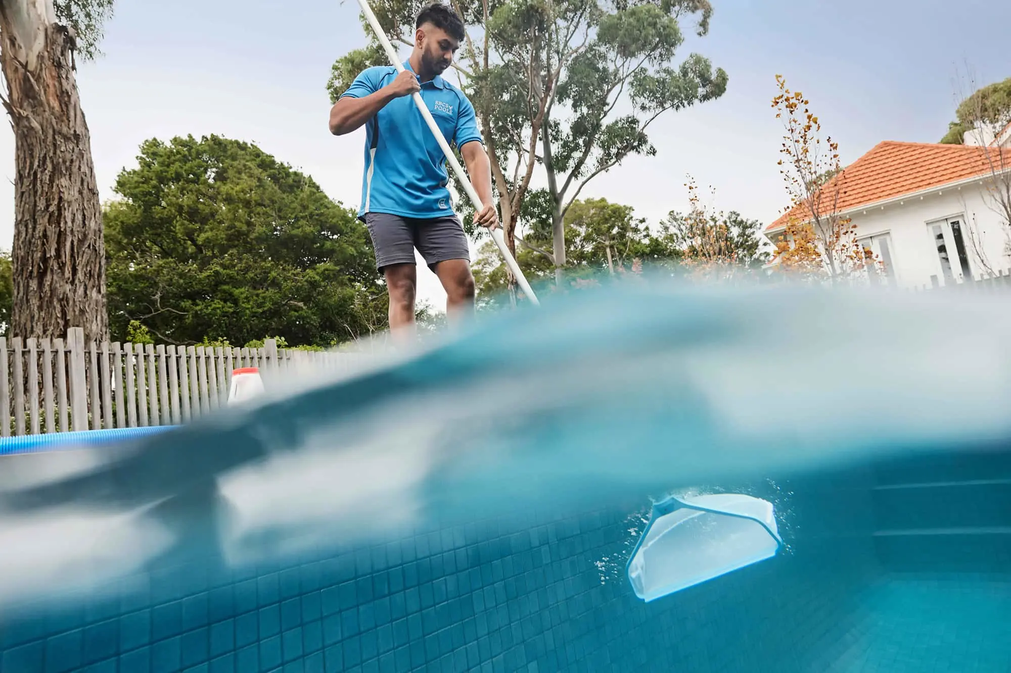 Pool maintenance worker in blue uniform using a long pole to clean a swimming pool, with a skimmer visible underwater.