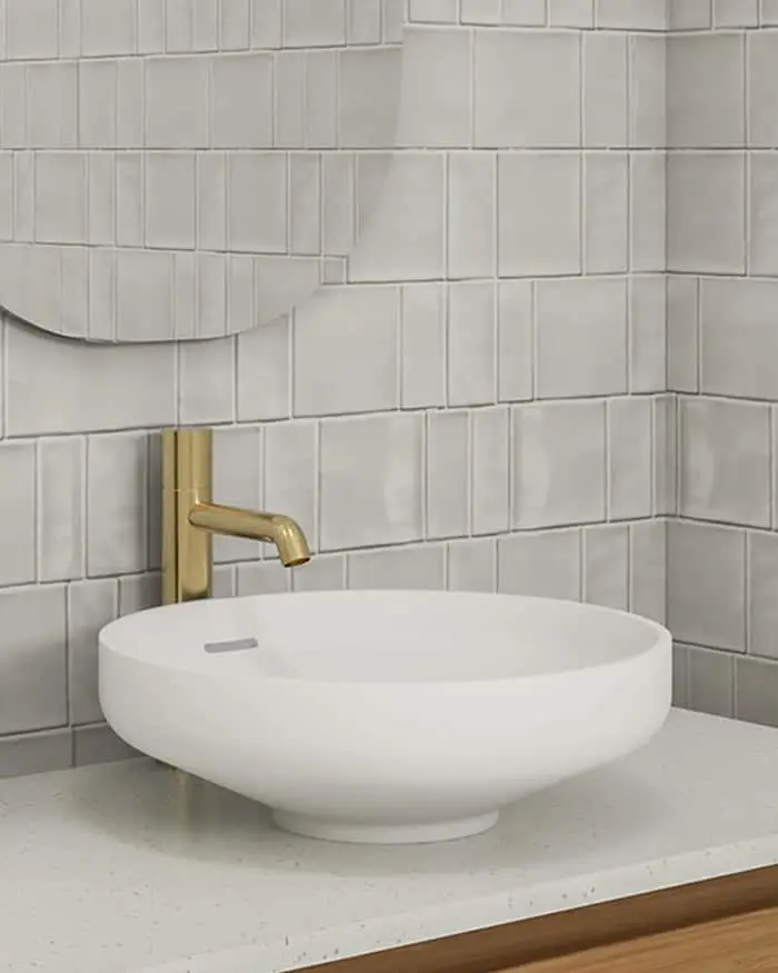 Modern bathroom with white vessel sink, brass faucet, and light gray vertical tile backsplash with circular mirror.