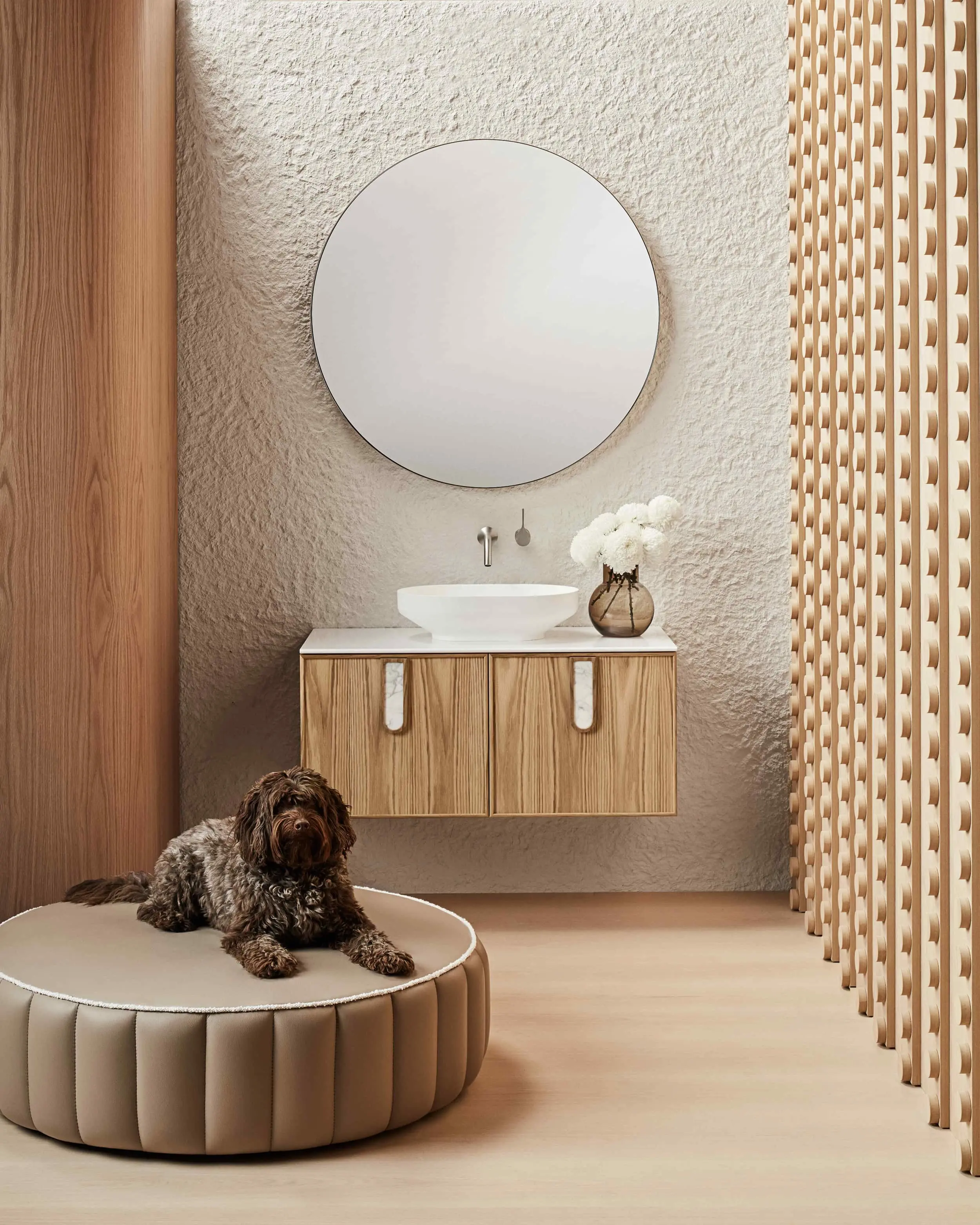 Modern bathroom with round mirror, wooden vanity, white sink, and brown dog resting on circular beige ottoman.