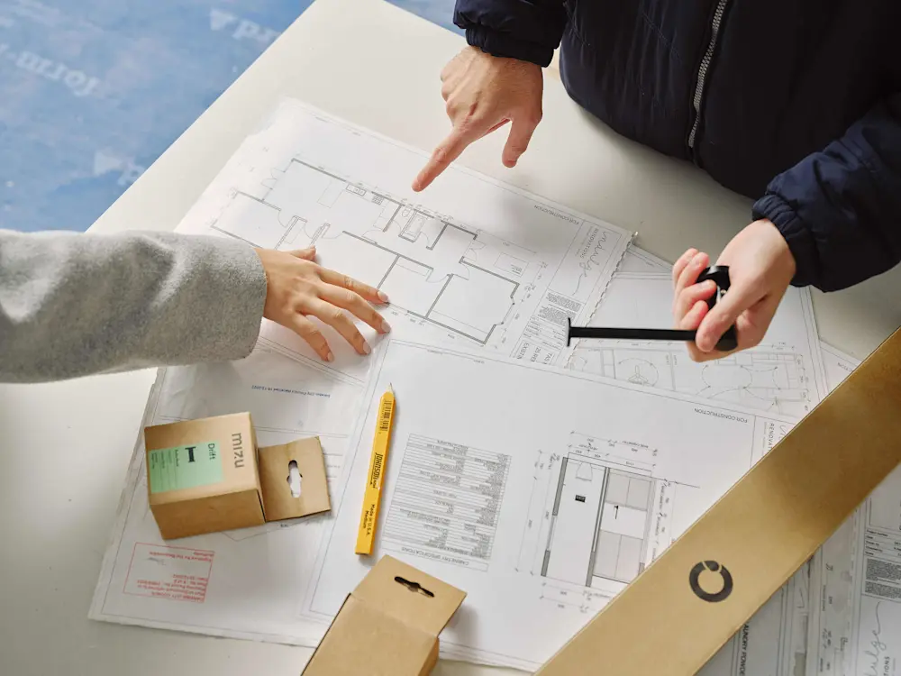 Two people reviewing architectural floor plans on a table with a pencil and small packaging boxes nearby.