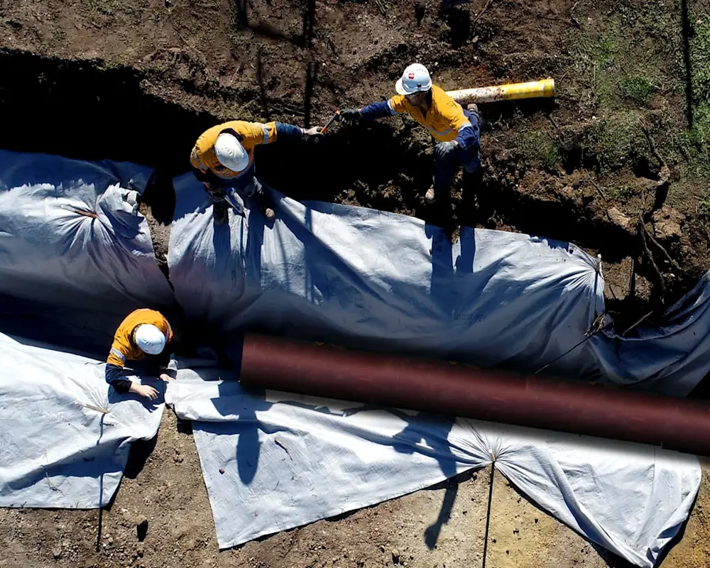 Construction workers in yellow safety gear installing large pipeline wrapped in protective white material in a trench.