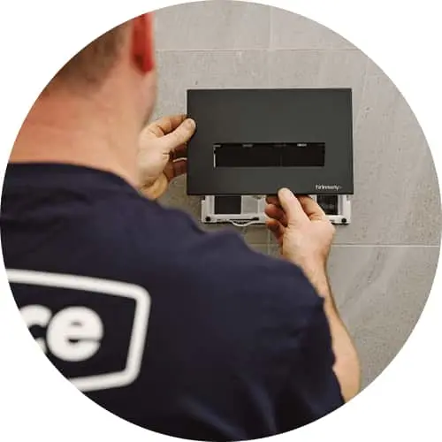 Person in blue uniform installing a black wall-mounted control panel or thermostat on a gray tiled wall.