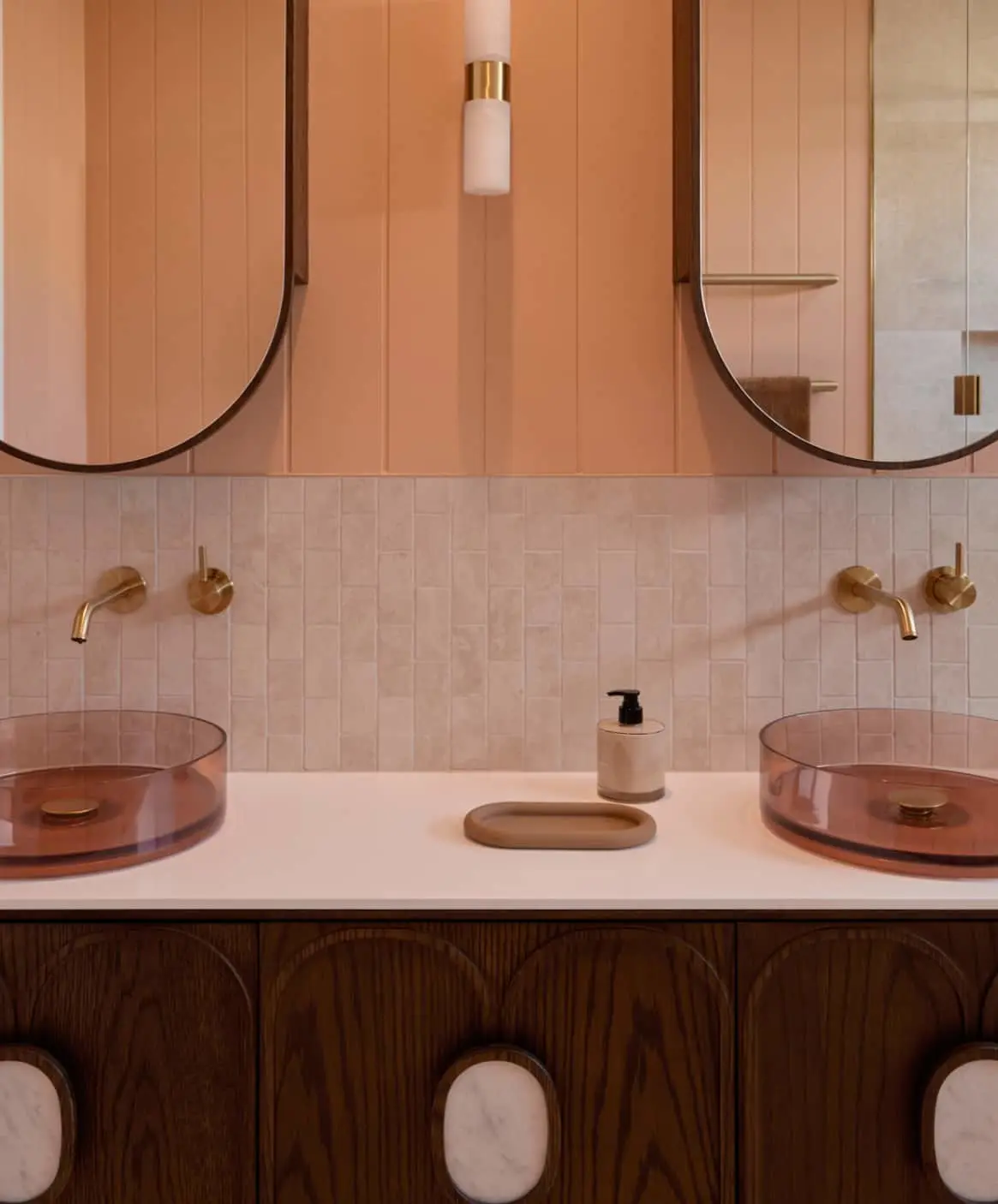 Modern bathroom with pink walls, brass fixtures, round mirrors, and amber glass sinks on a white countertop with wooden cabinetry.