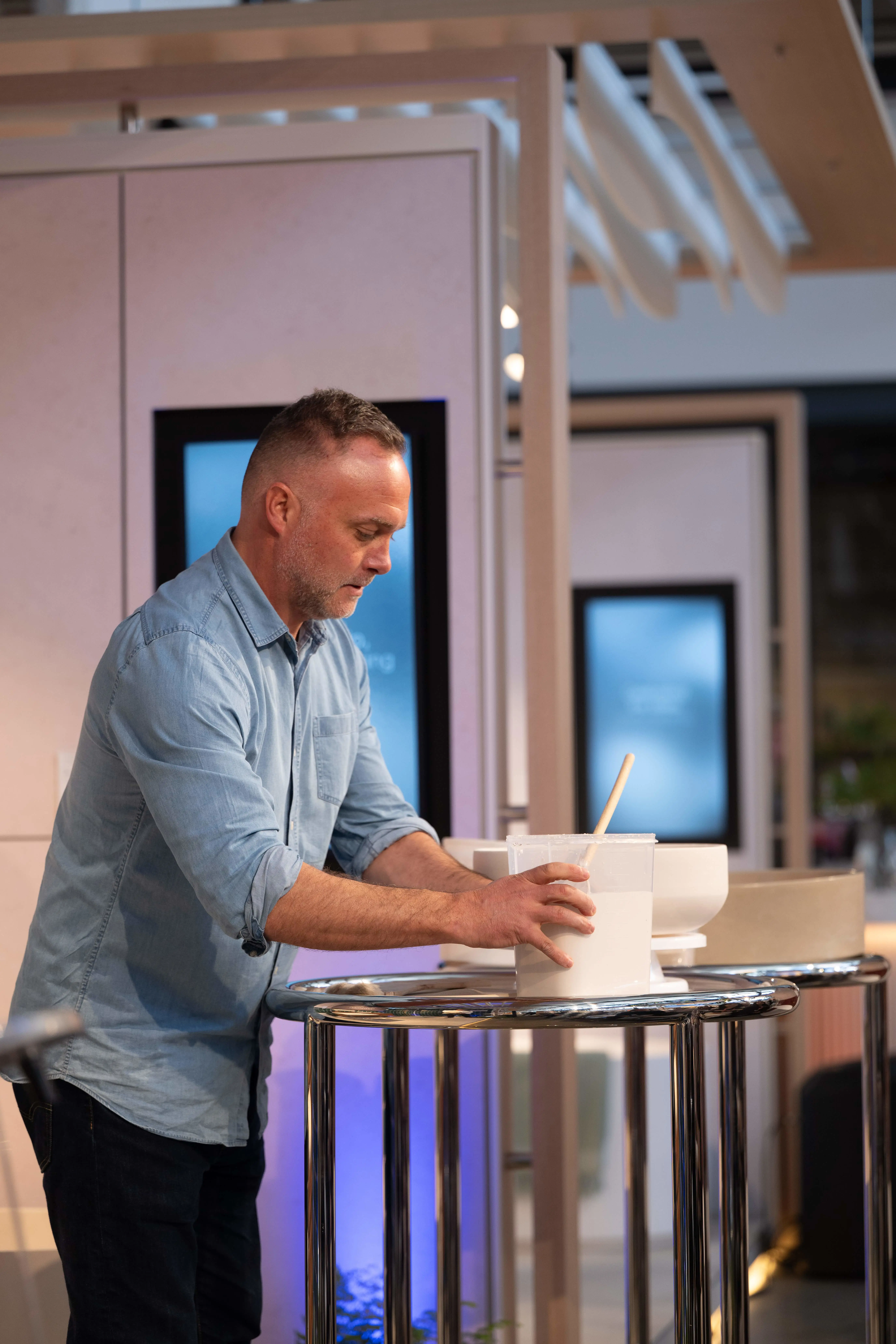 Man in light blue shirt shaping pottery on a wheel with hands, white clay bowl and tools visible on work surface.
