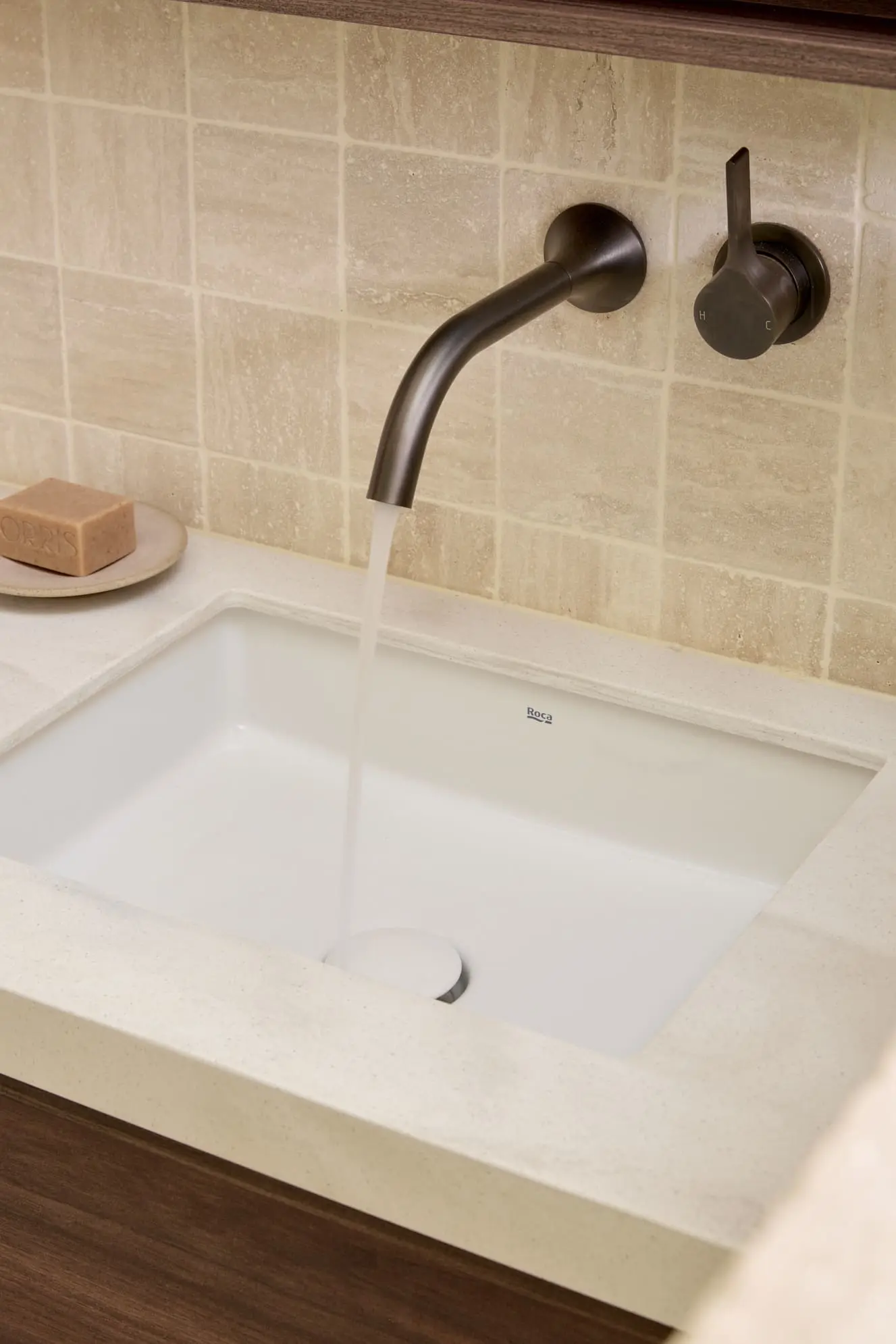Modern bathroom sink with wall-mounted bronze faucet running water, beige tile backsplash, and soap bar on counter.