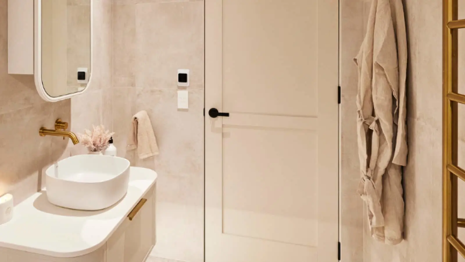 Modern beige bathroom with white vessel sink, brass fixtures, mirror, white door, and hanging beige robes.