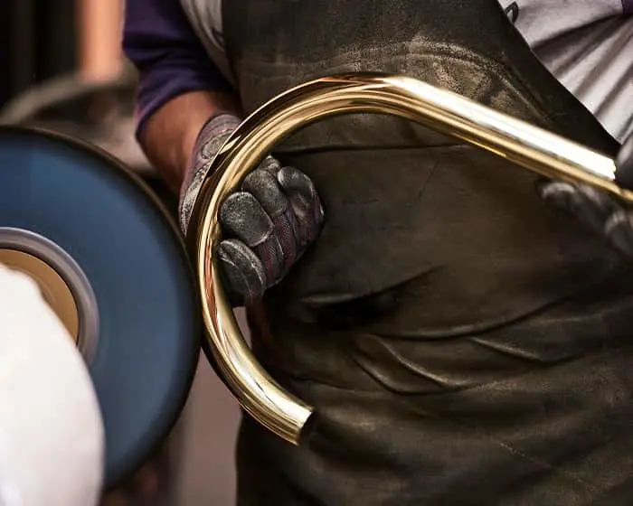 Craftsperson in protective gloves polishing a curved brass piece on a grinding wheel, showing metalworking craftsmanship.