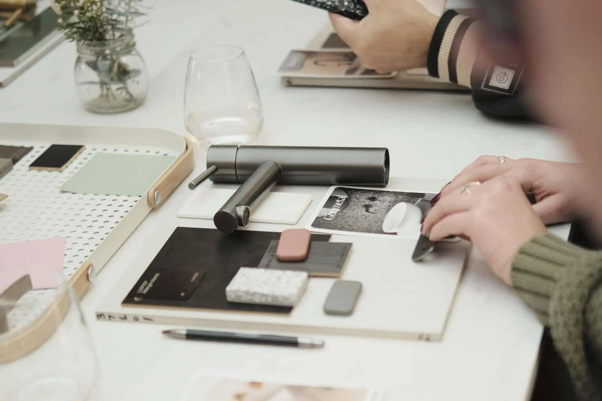 Hands arranging material samples on a white table with design tools, notebooks, and a small plant in the background.