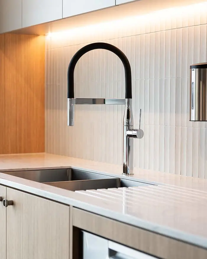 Modern kitchen sink with black and chrome pull-down faucet against white tile backsplash and light wood cabinetry.