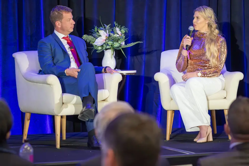 Two speakers on stage at an event, person in gold sequin top speaking into microphone while person in blue suit listens.