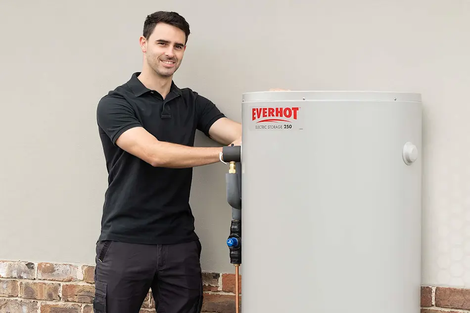 Person in black polo shirt installing or servicing an Everhot Electric Storage 250 water heater against beige wall.