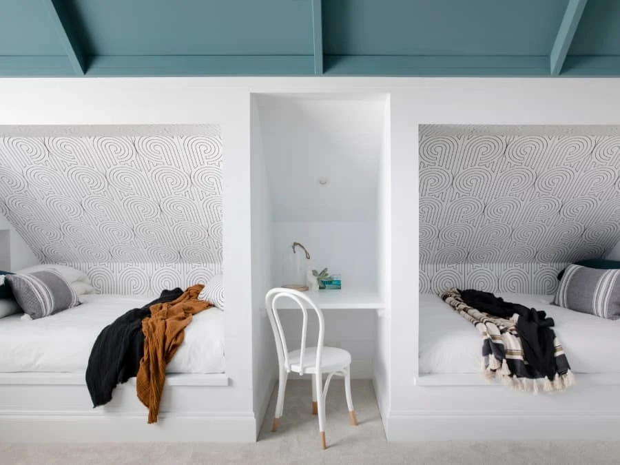 Modern bedroom with twin beds separated by small desk nook, patterned wallpaper, and teal ceiling beams.