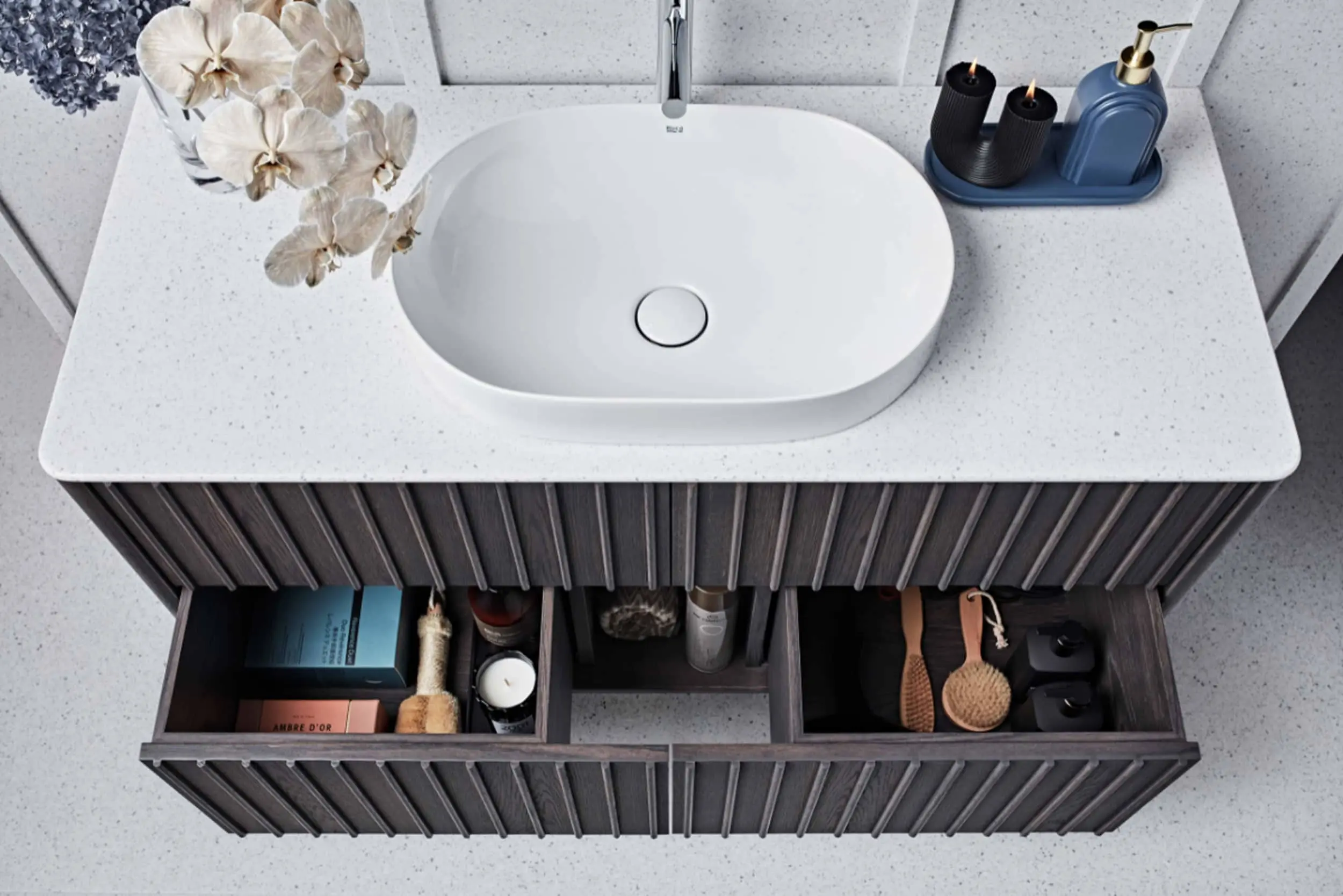Modern bathroom vanity with white oval sink, gray cabinet with open drawers containing toiletries, orchids and blue soap dispenser.