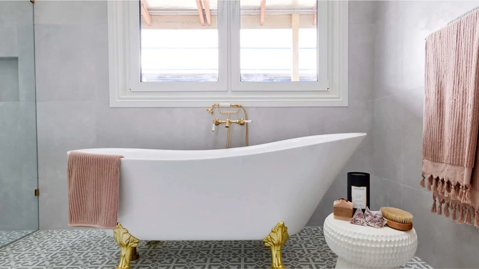 Modern bathroom with white clawfoot tub, gold fixtures, patterned floor tiles, and pink towels under bright windows.