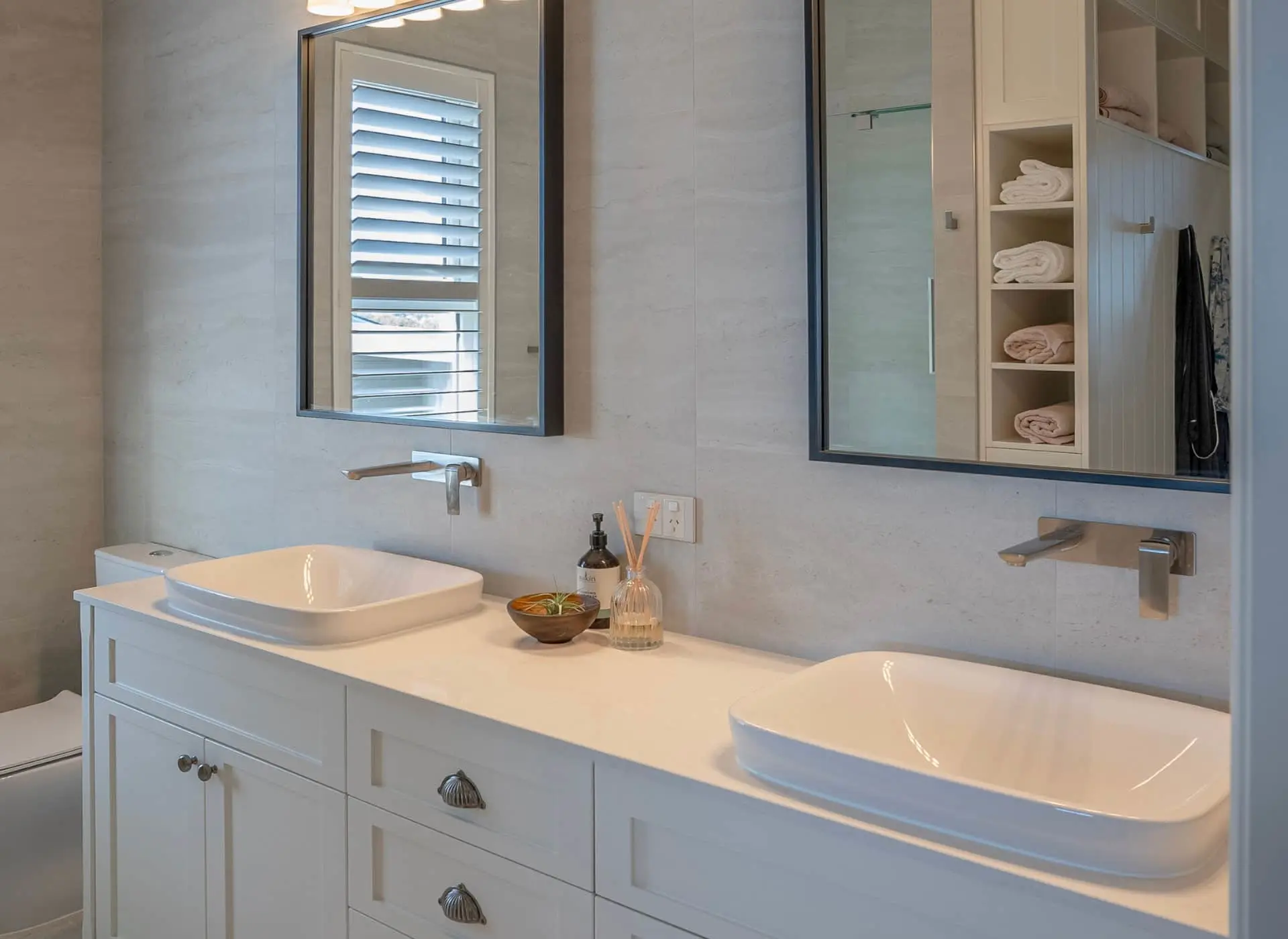 Modern bathroom with double white vessel sinks, white vanity, two mirrors, and built-in towel storage.