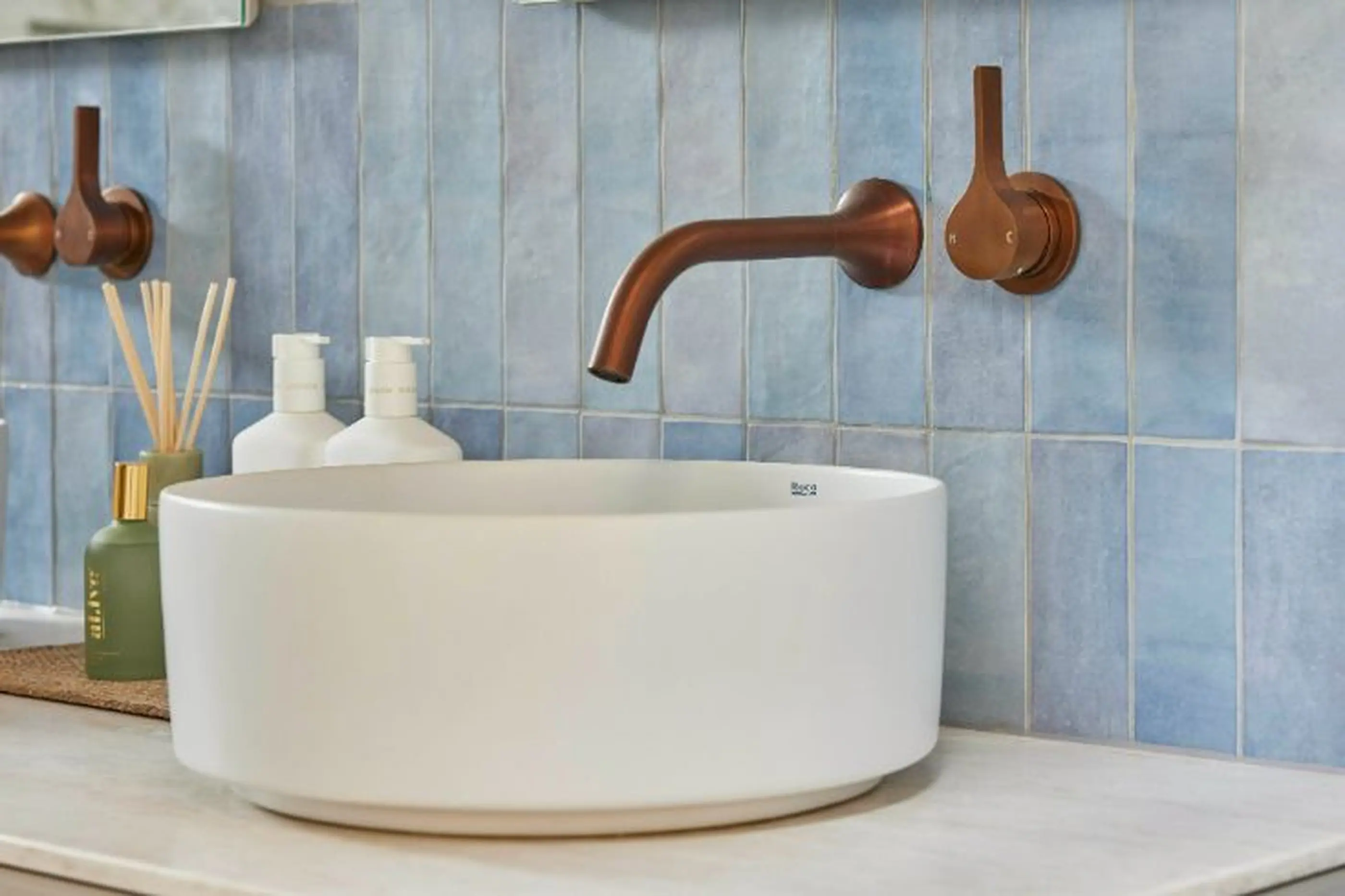 Modern bathroom sink with white vessel basin, copper wall-mounted faucet, and light blue vertical tile backsplash.