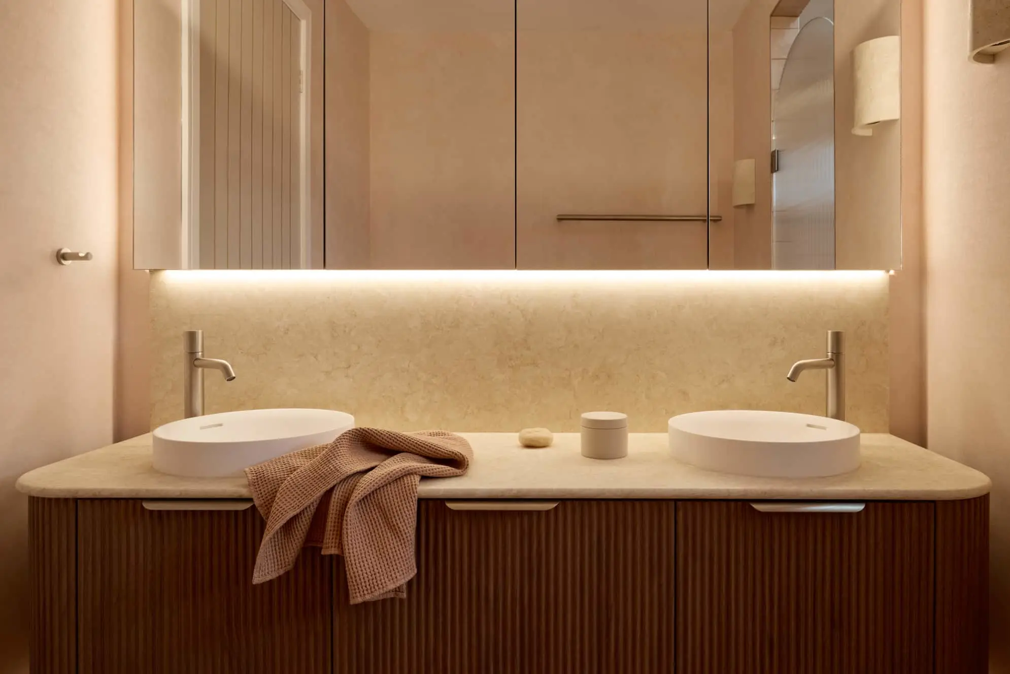 Modern bathroom with dual white vessel sinks on wooden vanity, beige countertop, and backlit mirror.