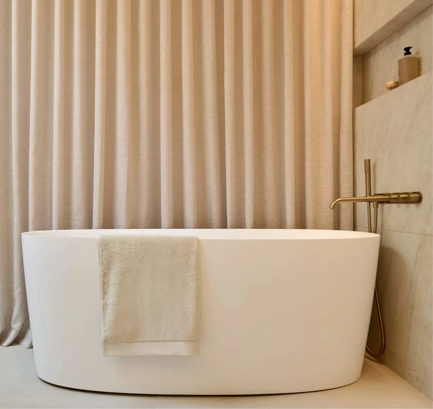 White oval freestanding bathtub with a cream towel draped over the side, brass wall-mounted faucet, and beige curtains.