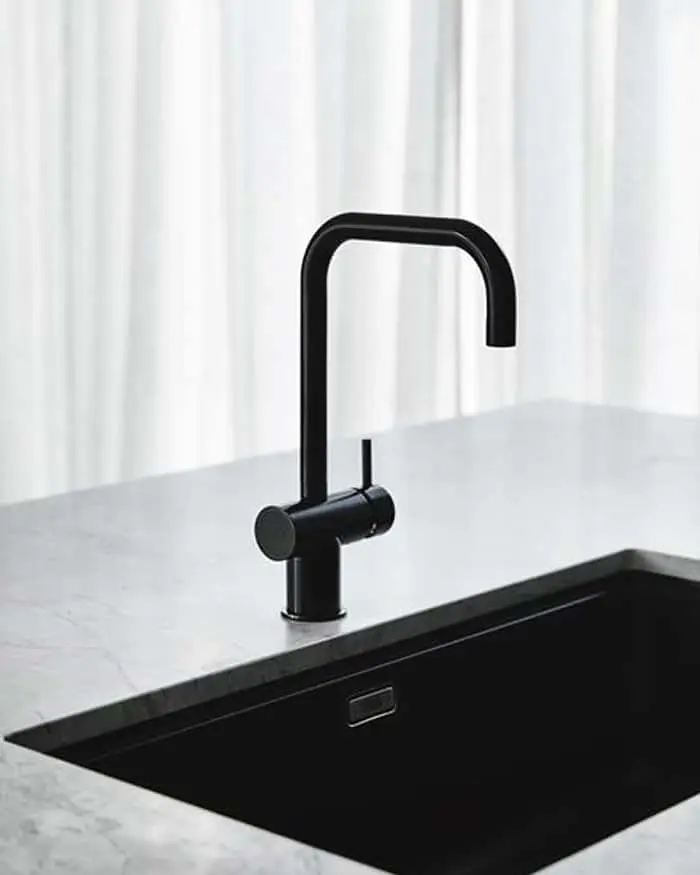 Modern black faucet with square spout mounted on white marble countertop with black sink against white curtains.