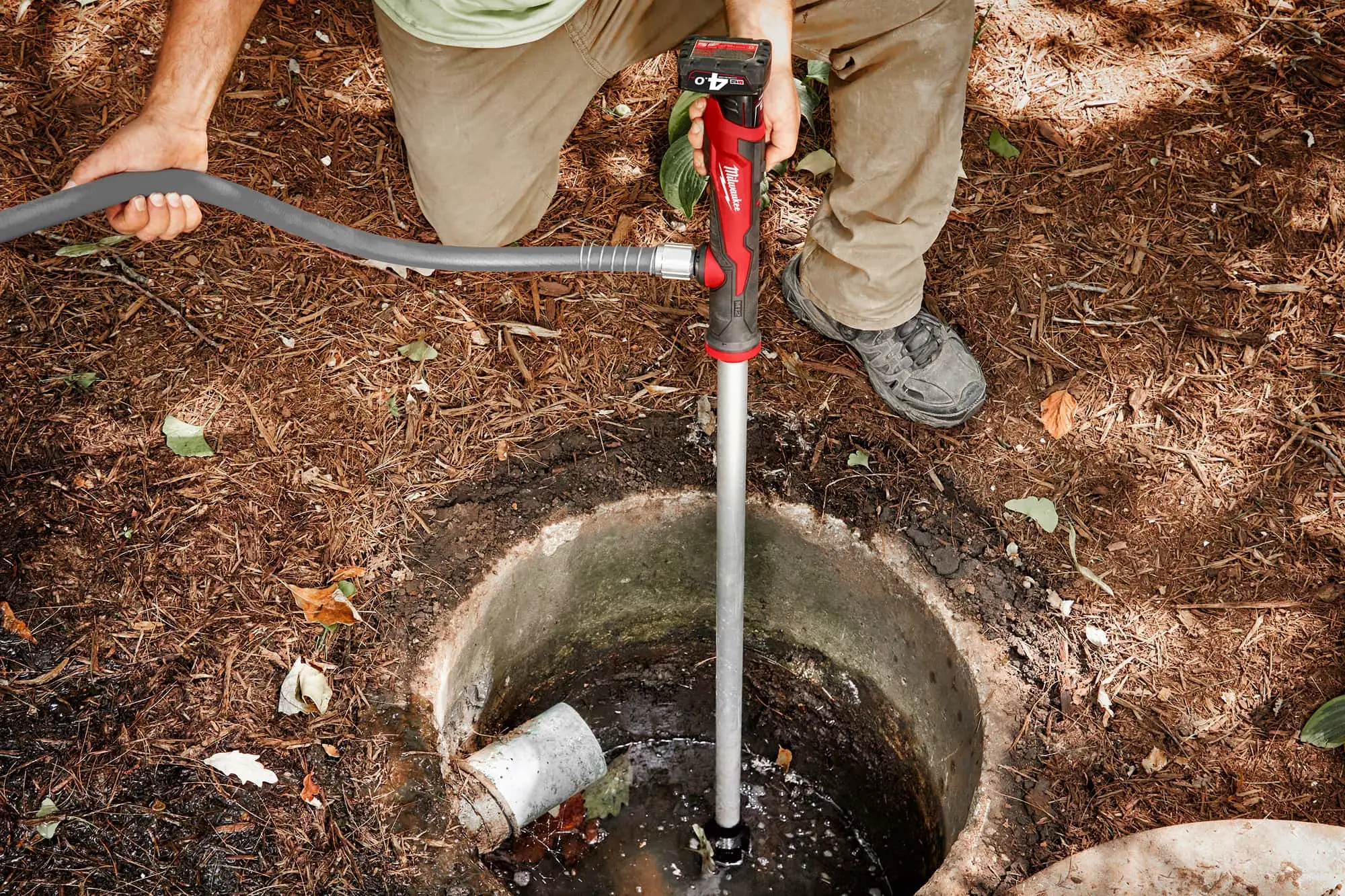 Person using a red Milwaukee power tool with extension to access water in an underground pipe or well on mulched ground.