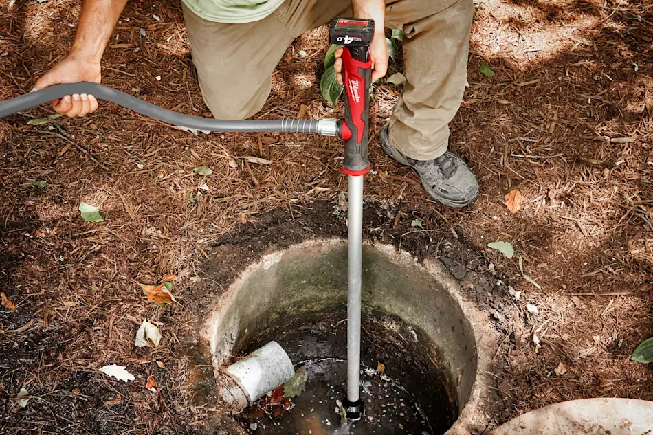 Person using a red Milwaukee power tool with extension to access water in an underground pipe or well on mulched ground.