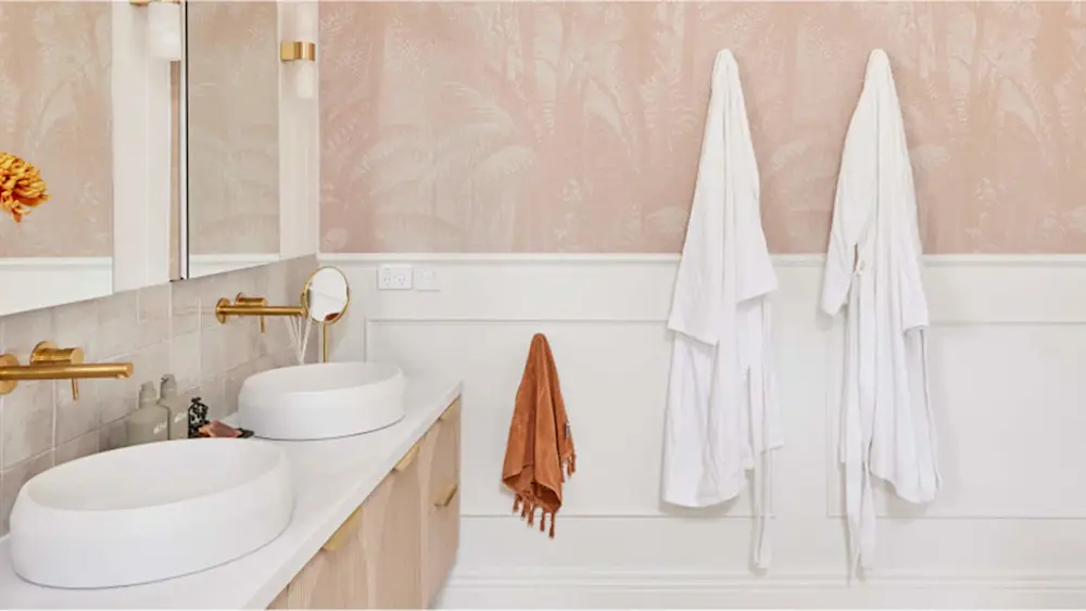 Elegant bathroom with pink floral wallpaper, double white sinks, gold fixtures, and white robes hanging on wall hooks.