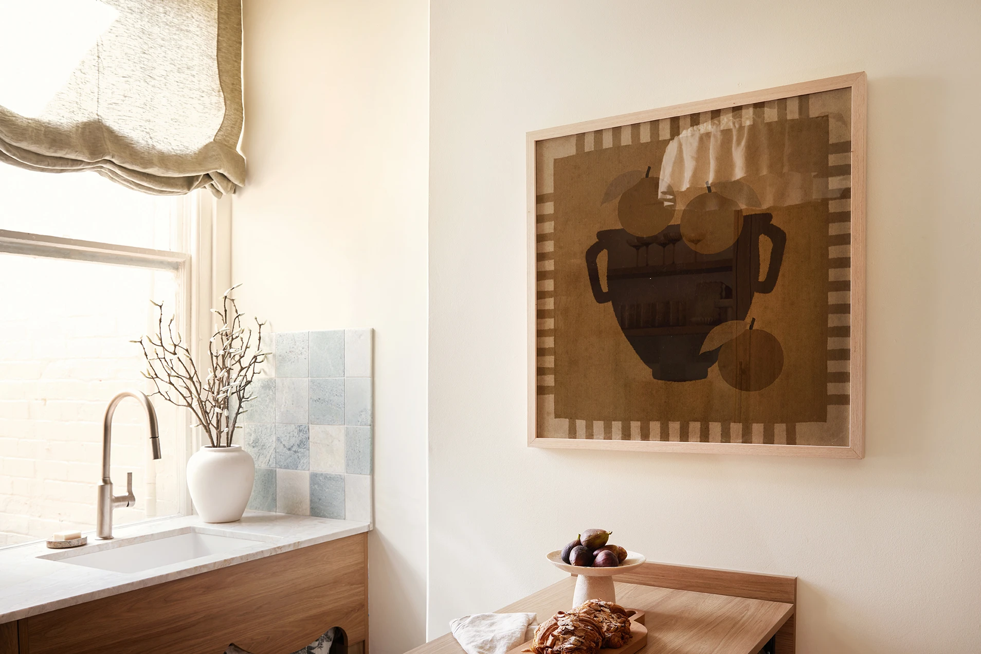Kitchen corner with a sink, blue tile backsplash, white vase with branches, and a framed still life artwork on the wall.