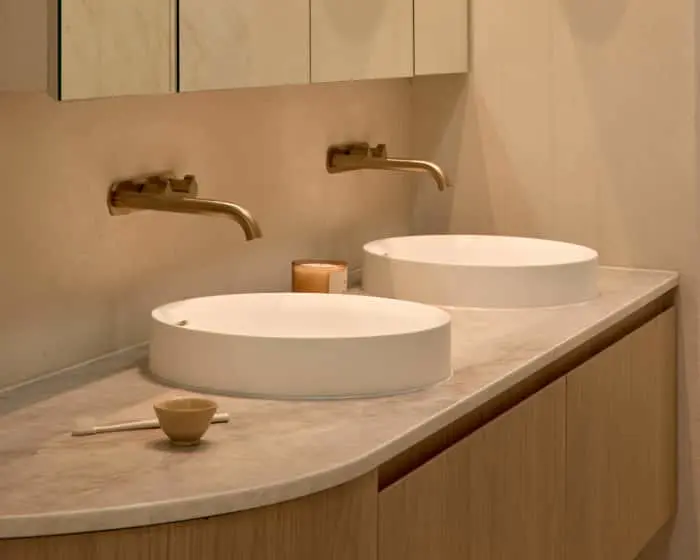Modern bathroom vanity with two white round vessel sinks, brass wall-mounted faucets, and light wood cabinetry.