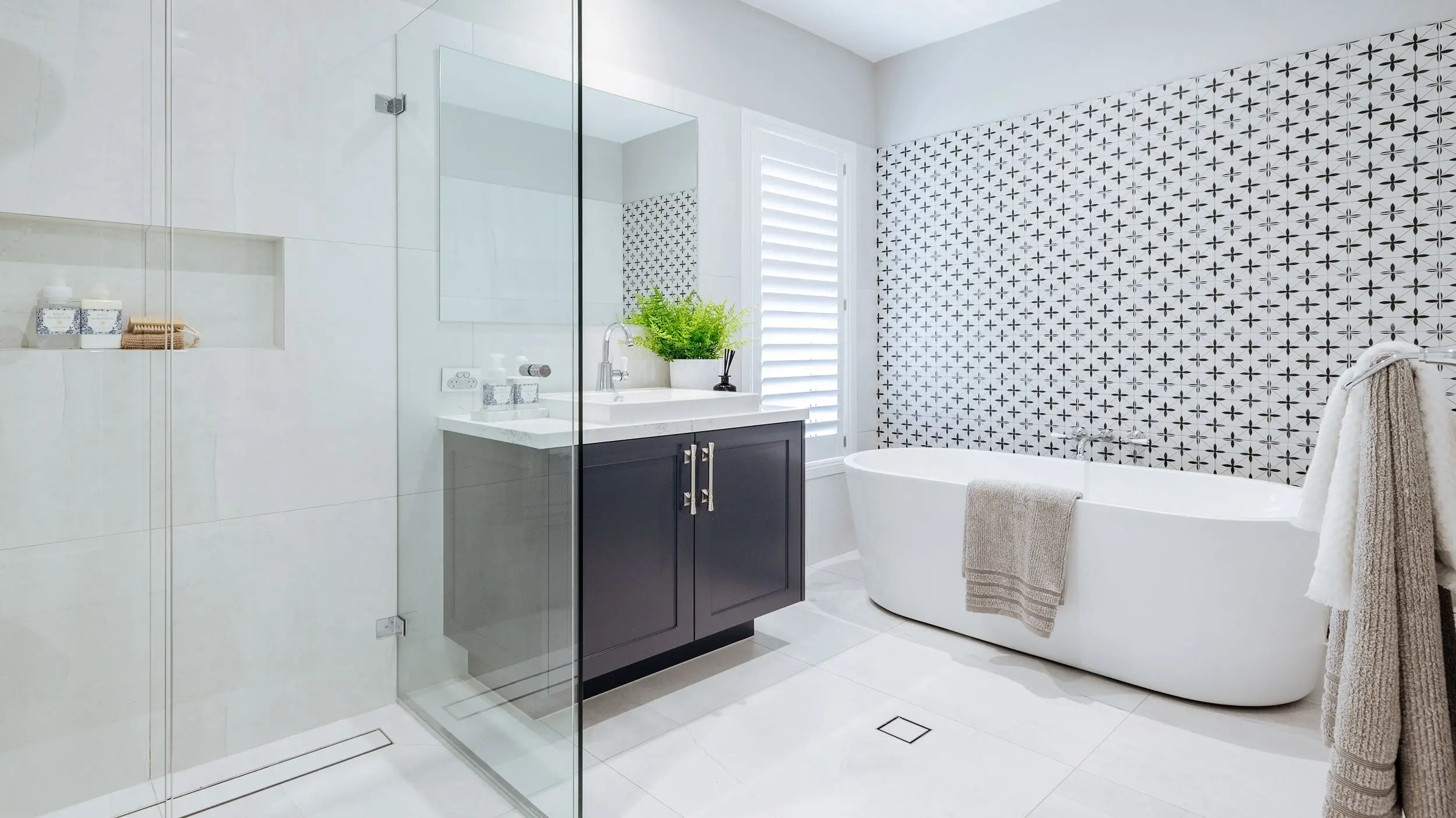 Modern bathroom with glass shower, dark vanity, white freestanding tub, and patterned accent wall.