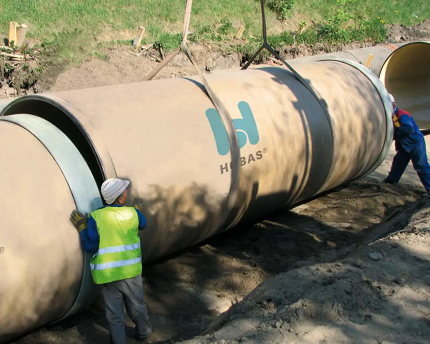 Workers installing large concrete drainage pipes with HOBAS branding at a construction site in a grassy area.