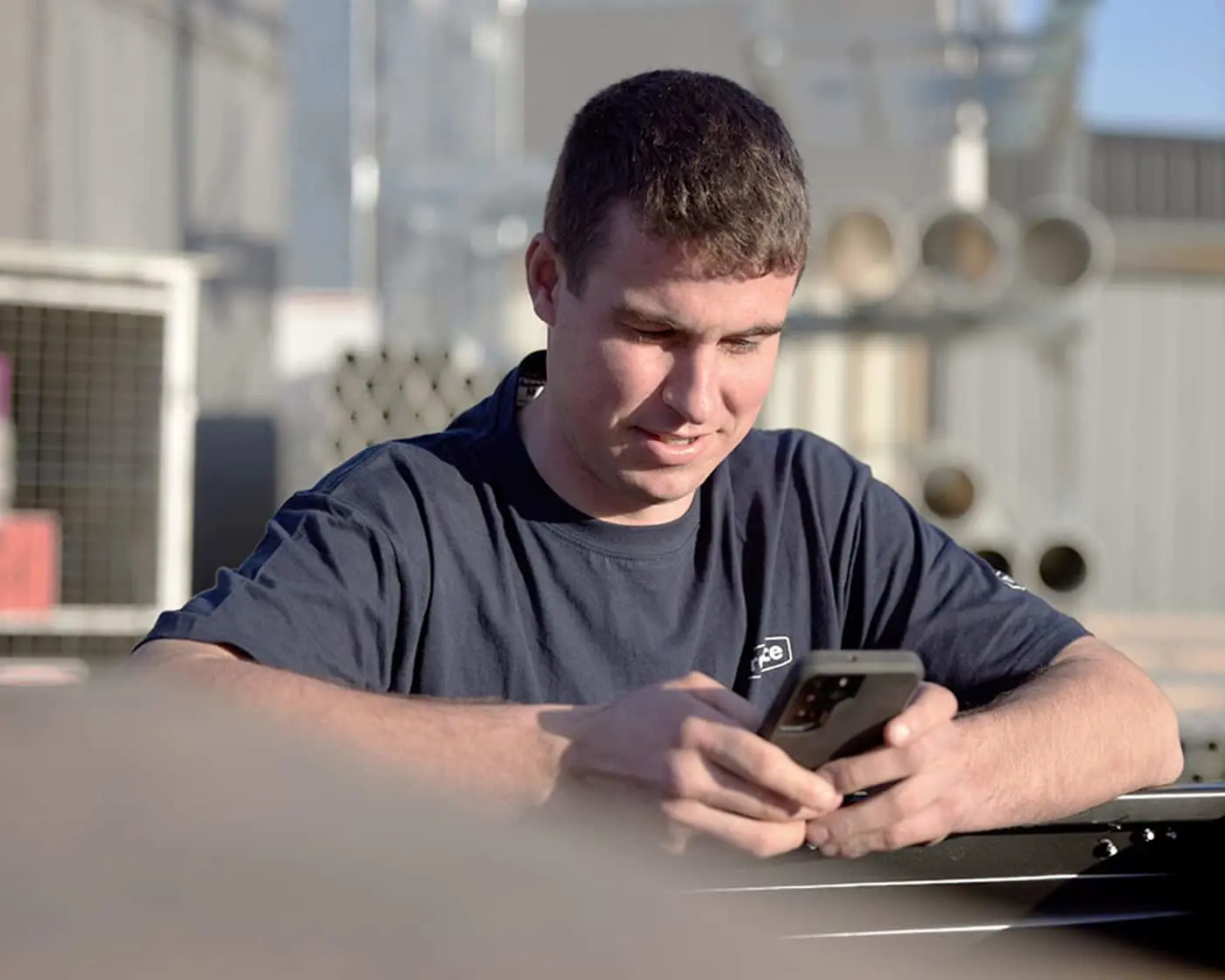 Person in navy blue t-shirt looking at smartphone in an industrial or workshop setting with blurred background.