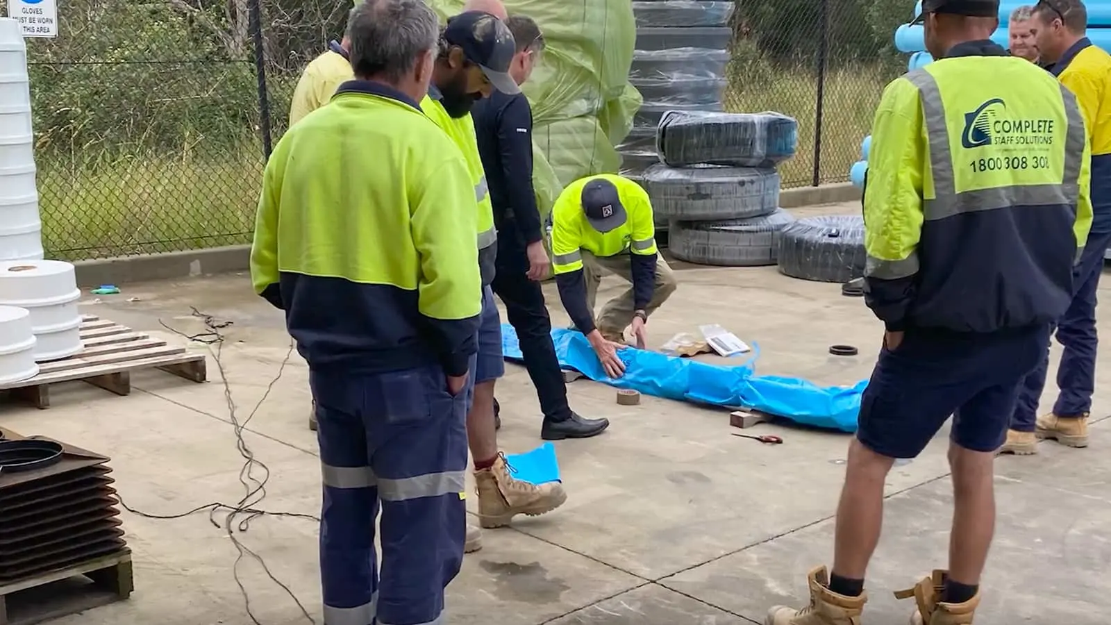 Construction workers in high-visibility gear examining blue material on concrete, with stacked supplies nearby.