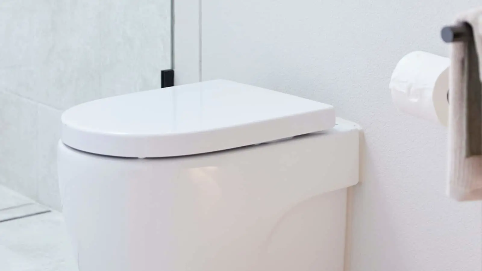 Modern white toilet with closed lid in a minimalist bathroom with toilet paper holder mounted on the wall.