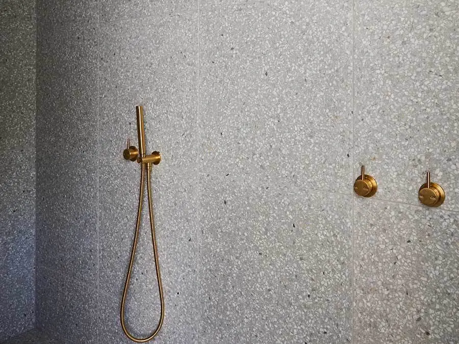 Modern shower with brass fixtures including handheld showerhead and control knobs against gray terrazzo wall.