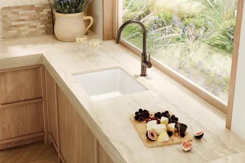 Modern kitchen with marble countertop, white sink, cheese board with figs, and window overlooking purple flowers.