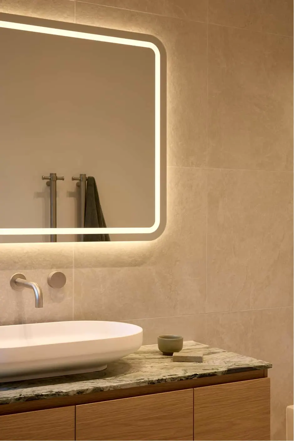 Modern bathroom with backlit mirror, white vessel sink on marble countertop, and beige tiled walls.