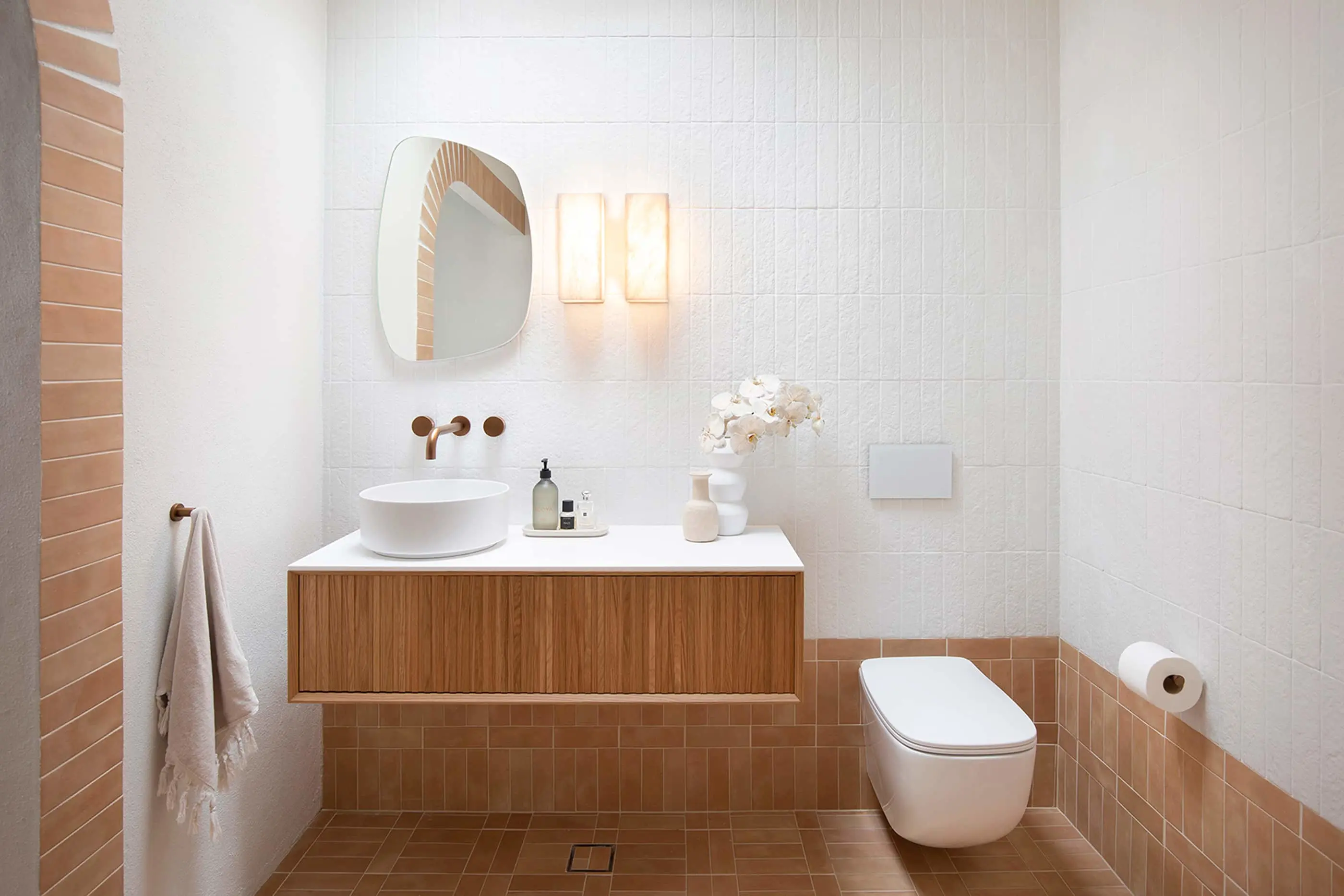 Modern bathroom with white walls, terracotta tiles, wooden vanity, white sink, wall-mounted toilet, and decorative flowers.