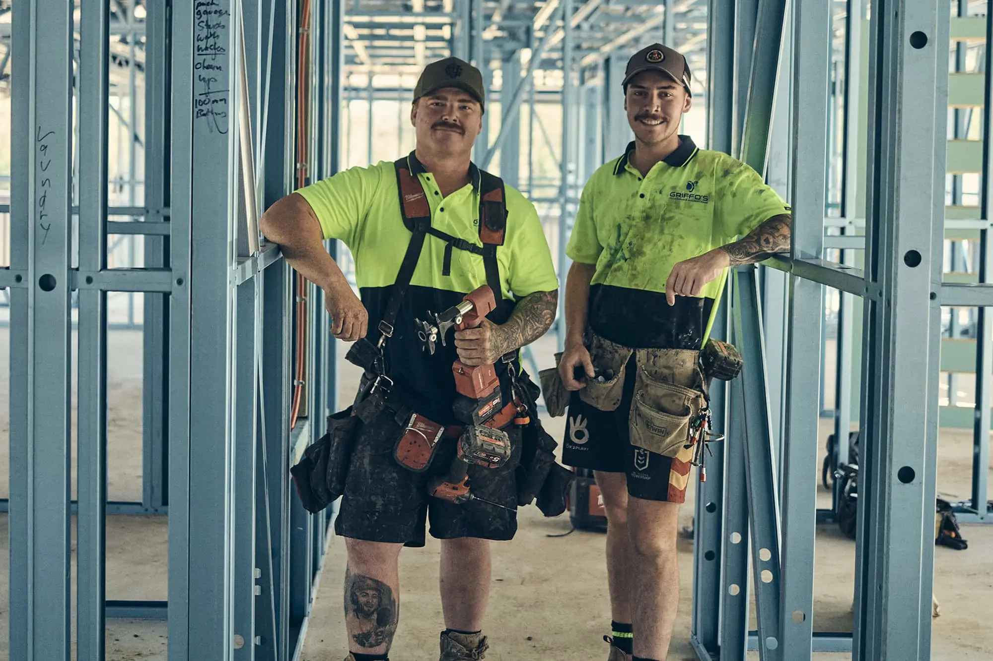 Two construction workers in bright yellow shirts and tool belts standing inside a metal framed building under construction.