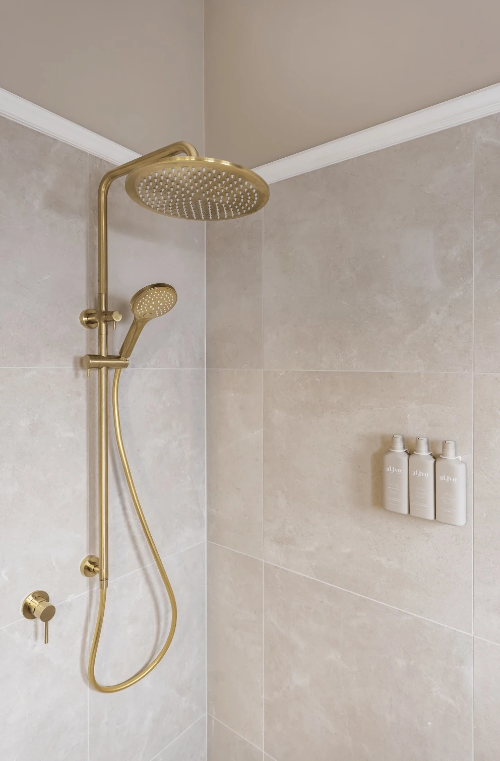 Modern brass rainfall shower system with handheld attachment mounted on beige tiled wall with three toiletry bottles.