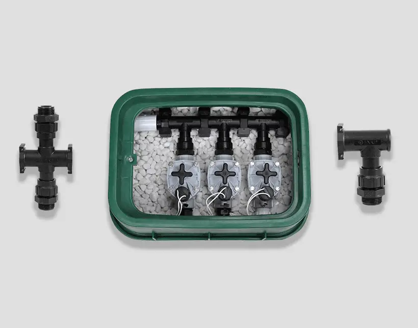 Green irrigation valve box with three control valves and white stones, flanked by black plastic pipe fittings.
