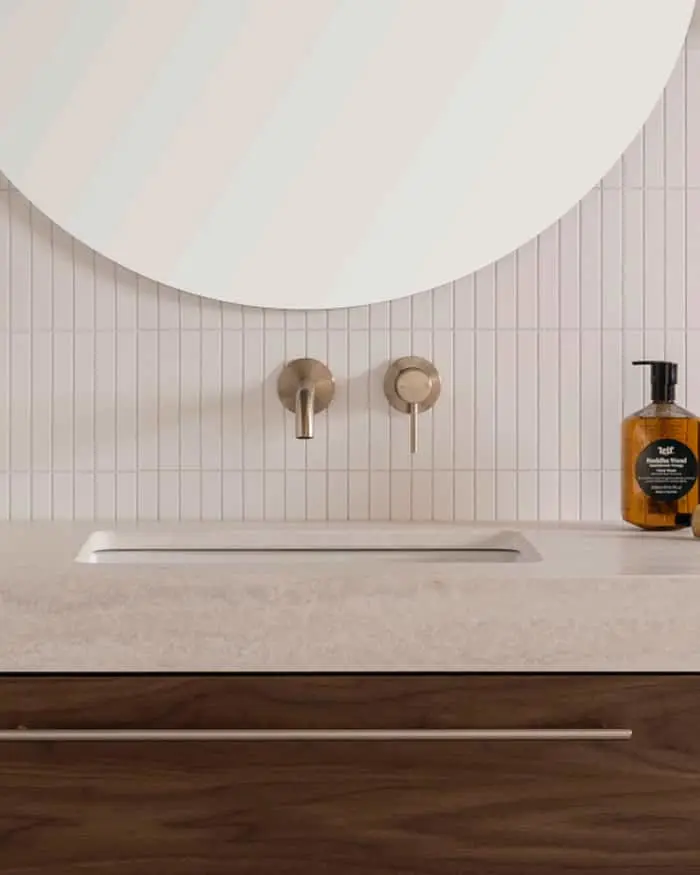 Modern bathroom sink with brass wall-mounted faucets, white tile backsplash, round mirror, and amber soap dispenser.