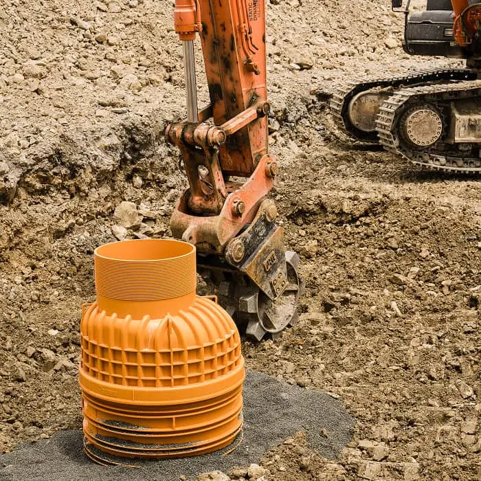 Orange excavator arm next to a plastic drainage pipe at a construction site with dirt and gravel ground.