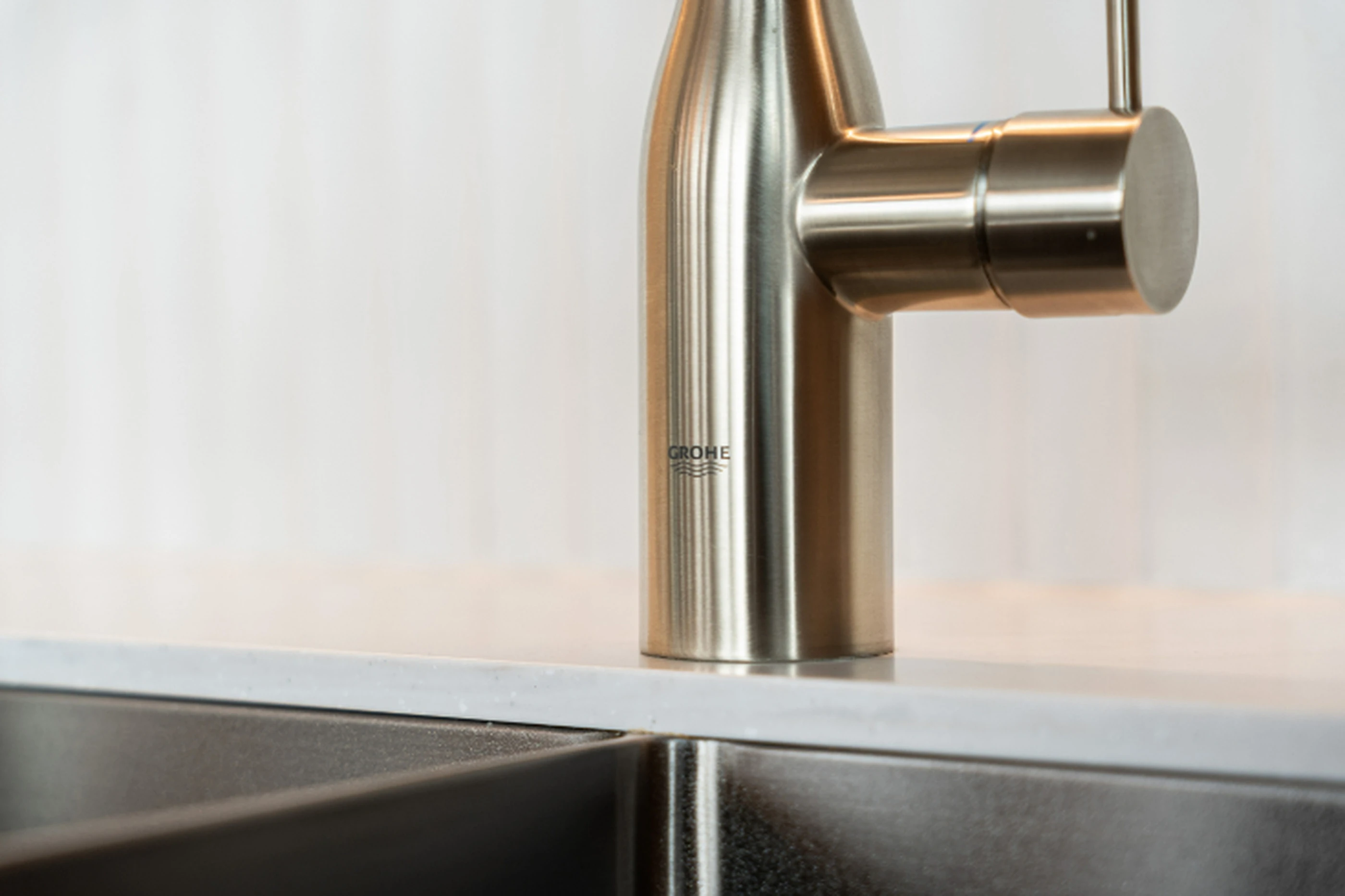 Close-up of a stainless steel GROHE faucet with cylindrical handle mounted on a white countertop above a dark sink.
