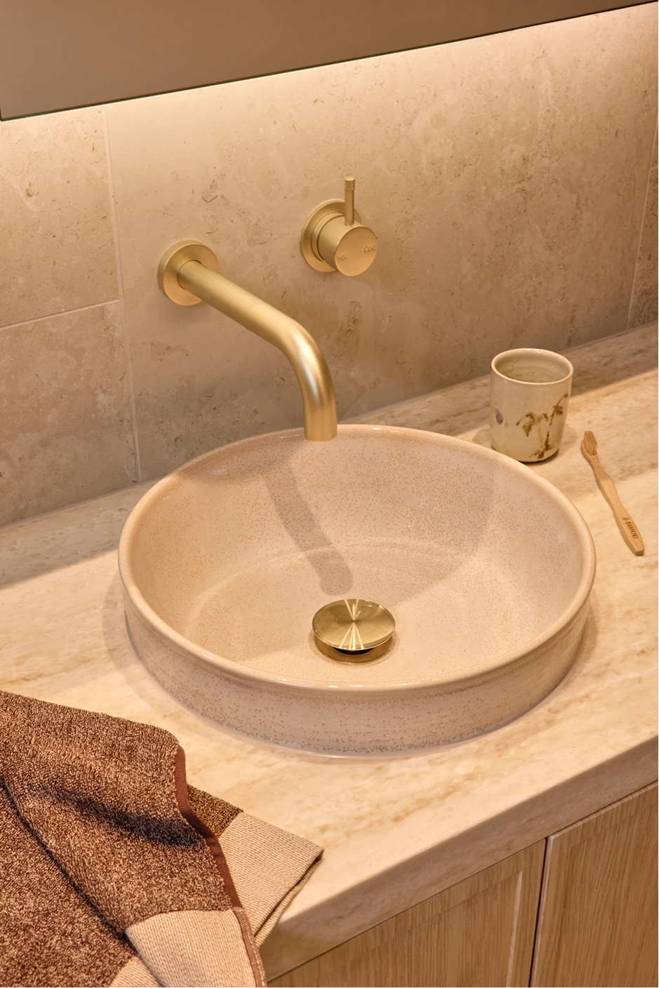 Round beige vessel sink with brass wall-mounted faucet on marble countertop with cup and toothbrush beside it.