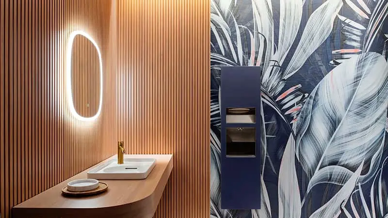 Modern bathroom with wood-paneled wall, illuminated oval mirror, and tropical leaf wallpaper in blue and white tones.